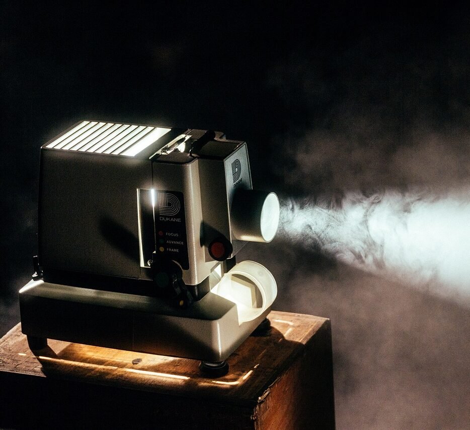 A vintage Dukane projector projecting a beam of light in a dimly lit room, sitting on a wooden platform with light reflecting off the projector.