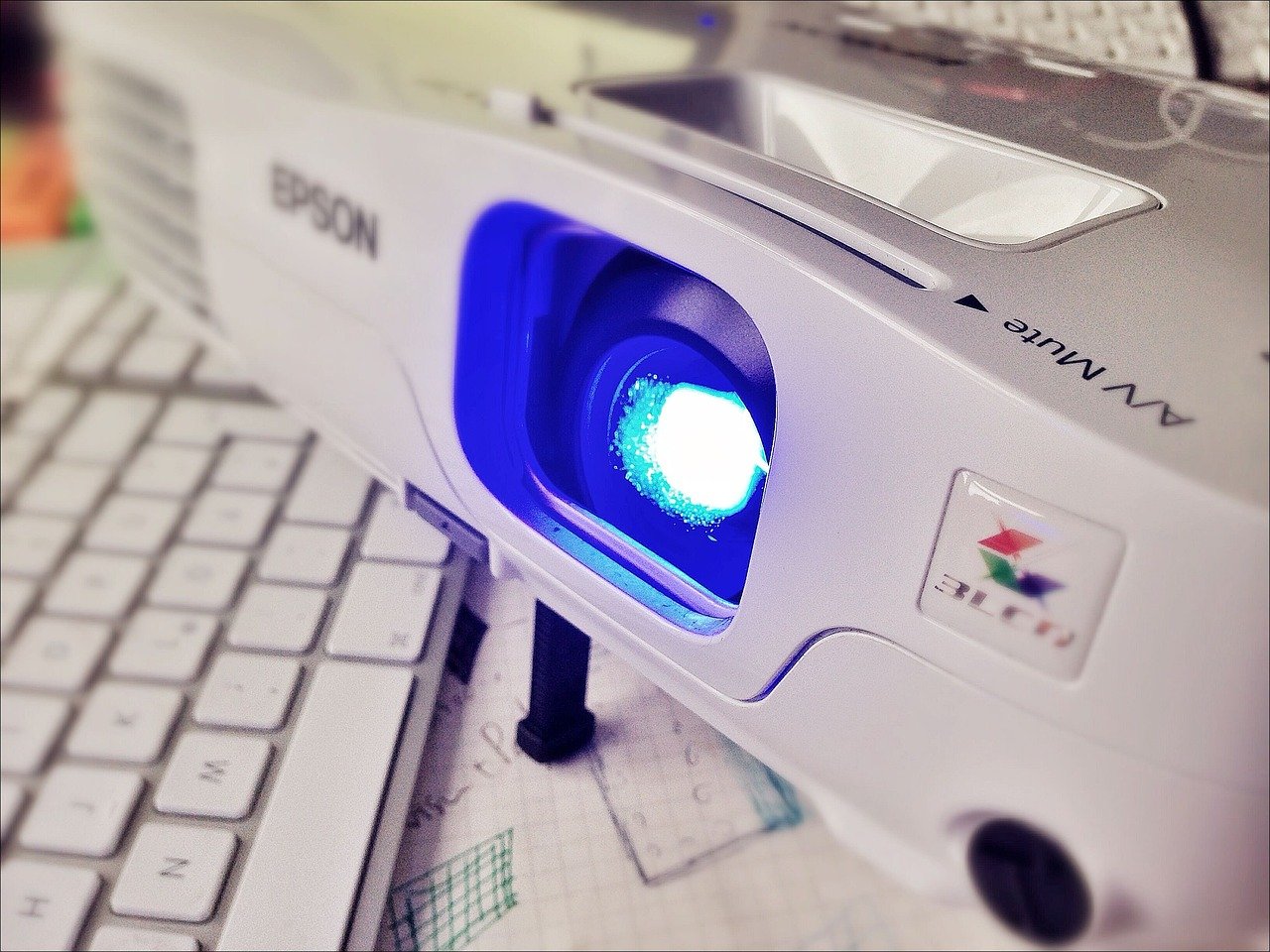 Close-up of an Epson projector with a bright blue lens light, placed next to a computer keyboard.